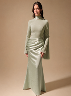 A woman modeling a floor-length sage green dress with a mock neck, long sleeves, and draped detailing at the waist, set against a solid beige studio background.