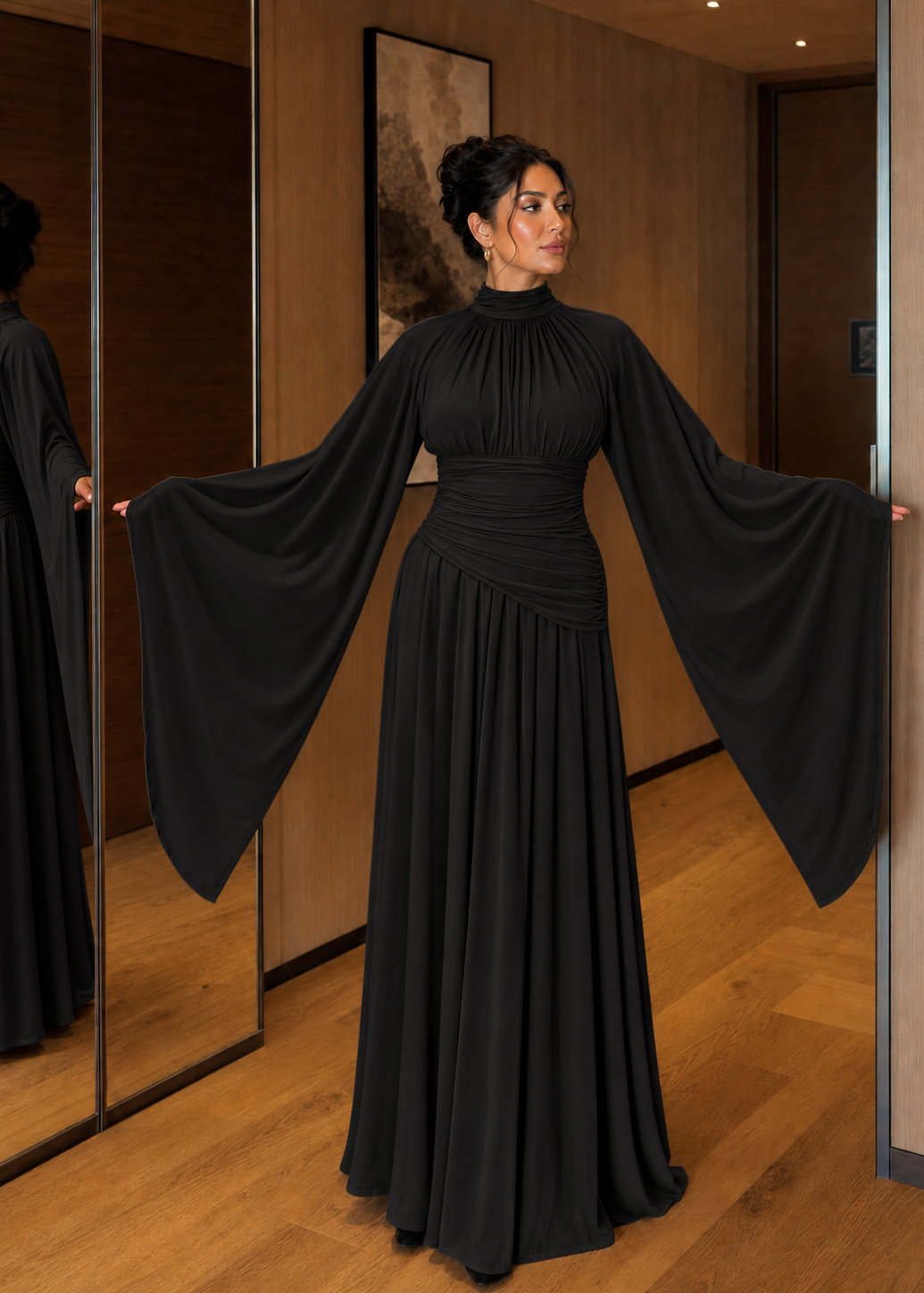 A woman wearing a floor-length black gown with dramatic wide sleeves and a gathered waist, standing in a warmly lit indoor hallway with wood walls and mirrors.
