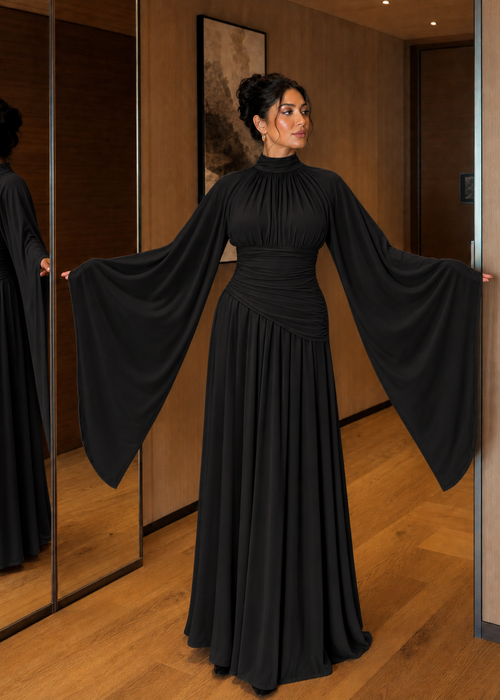 A woman wearing a floor-length black gown with dramatic wide sleeves and a gathered waist, standing in a warmly lit indoor hallway with wood walls and mirrors.