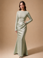 A woman modeling a sage green long-sleeve maxi dress with a gathered ruched bodice and a fitted mermaid-style skirt, posed against a solid warm beige studio backdrop.