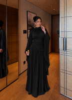 A woman wearing a floor-length black gown with a high mock neck, long draped sleeves, and a gathered waist, standing in a modern indoor hallway with wood flooring and mirrored walls.