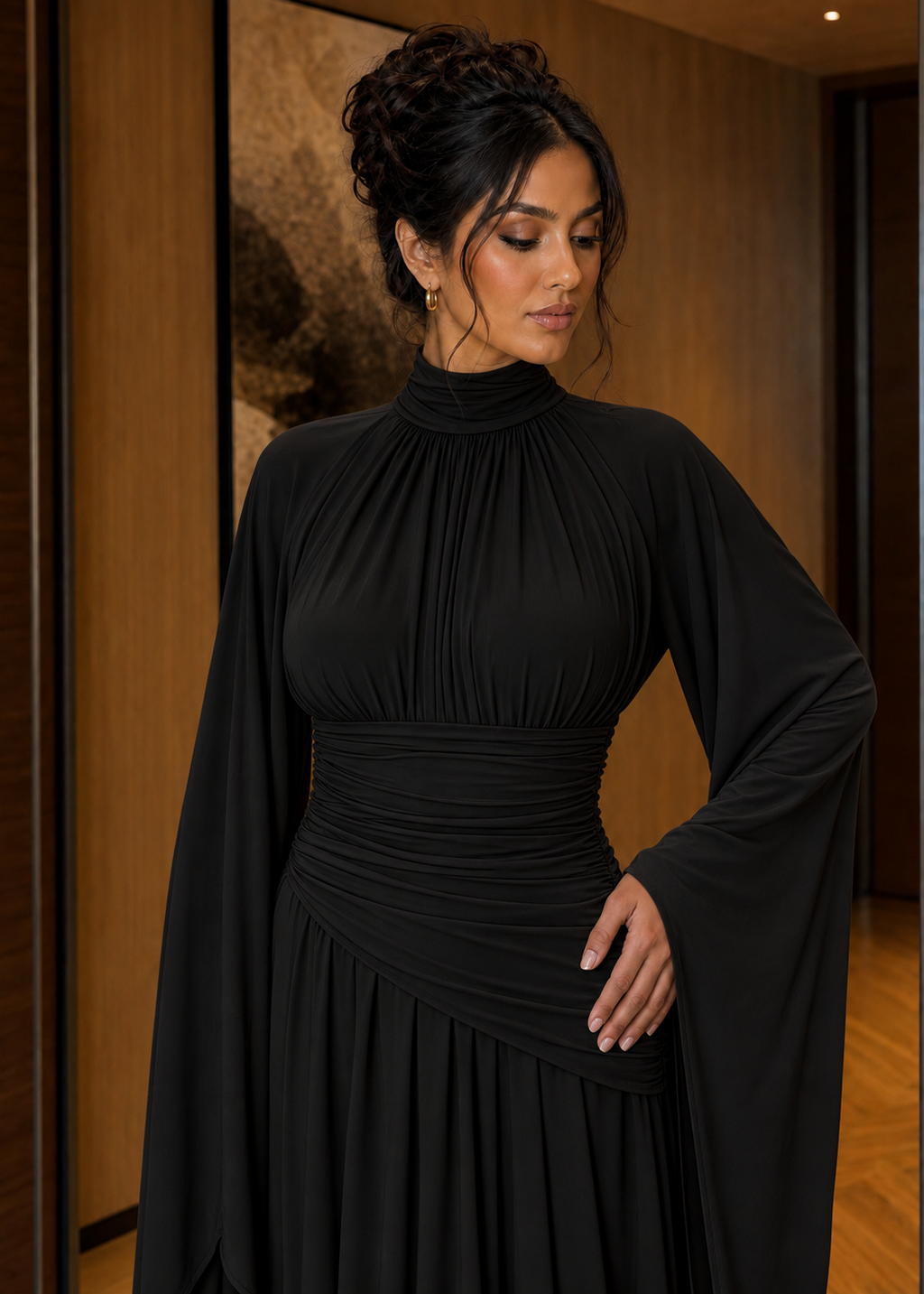 A woman wearing a black high-neck maxi dress with dramatic draped cape sleeves and a gathered waist, photographed indoors against a warm wood-paneled background.