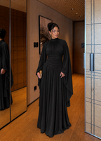 A woman wearing a floor-length black gown with draped sleeves and a gathered waist, standing in a modern indoor hallway with wood flooring and mirrors.
