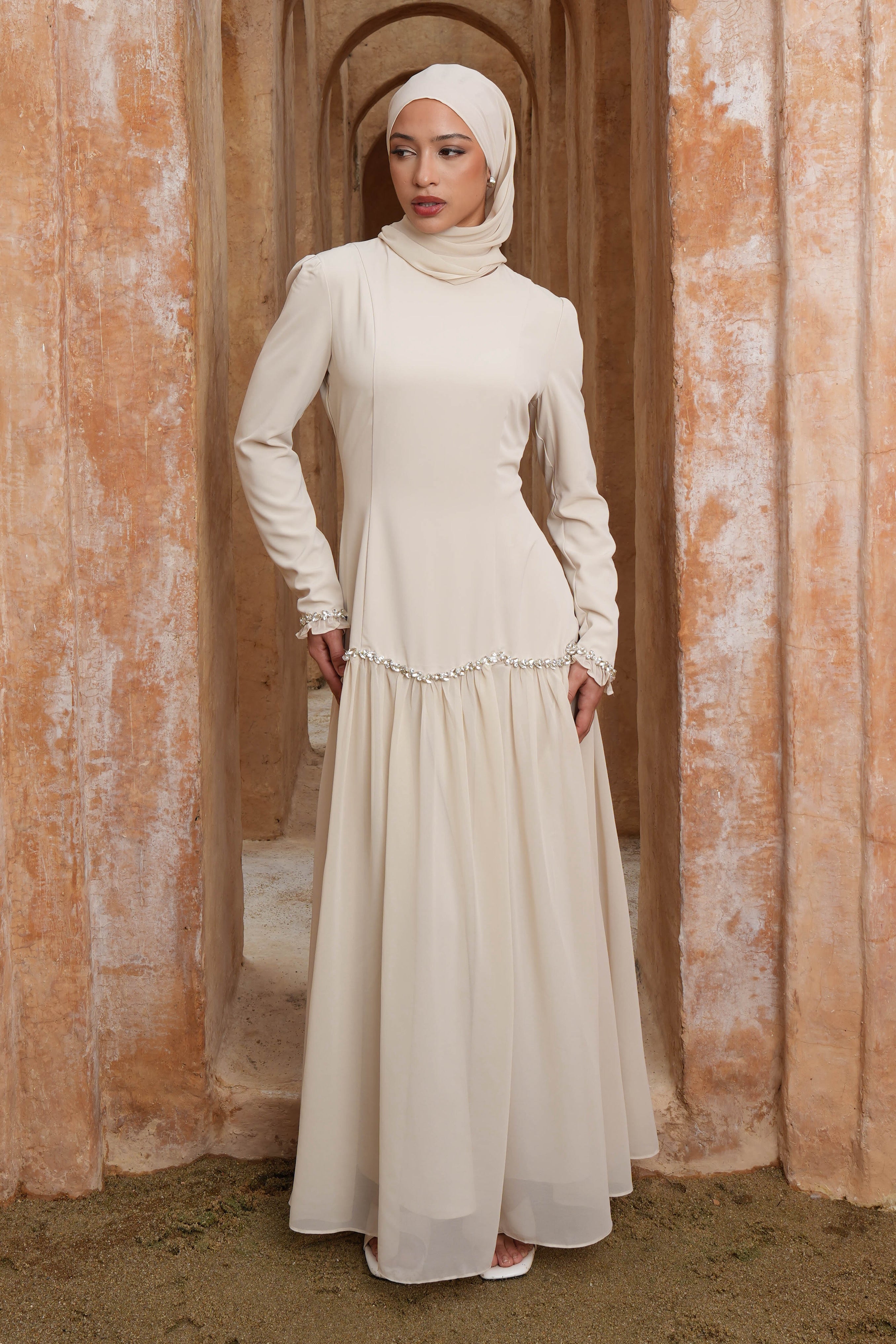 Woman in a long, light-colored dress standing in an arched doorway with a warm-toned wall.