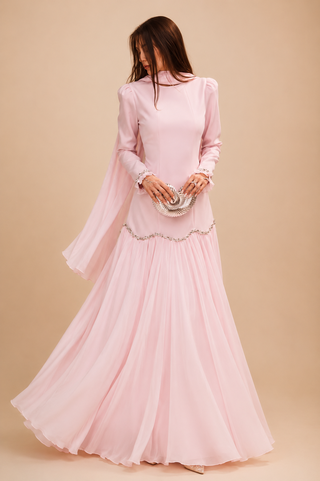A woman wearing a long-sleeved light pink maxi dress with a high neck, sheer draped sleeves, and a flared pleated skirt, holding a metallic clutch against a solid beige background.
