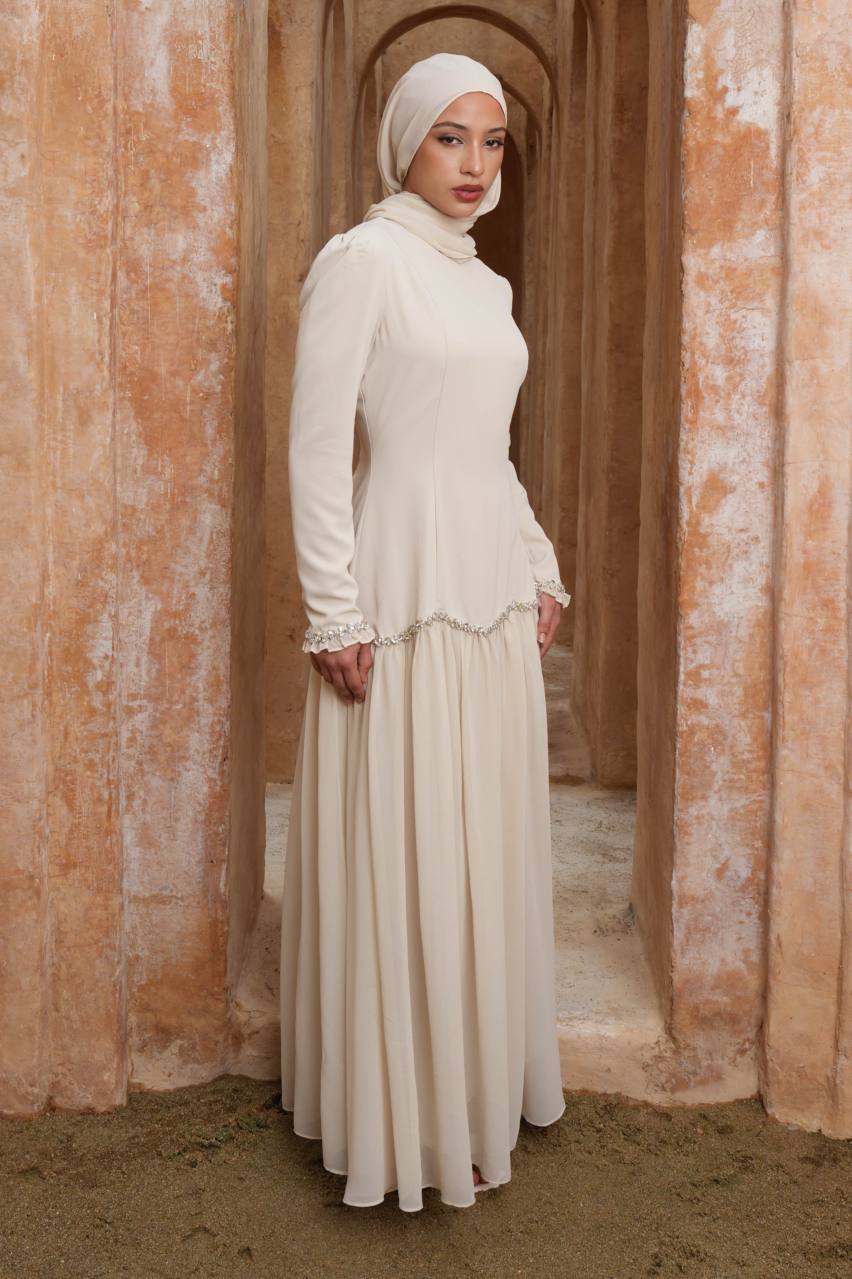 Woman in a long, light-colored dress standing in an arched doorway with warm-toned walls.