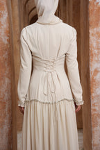Person wearing a long beige dress with a hood in front of a textured wall.