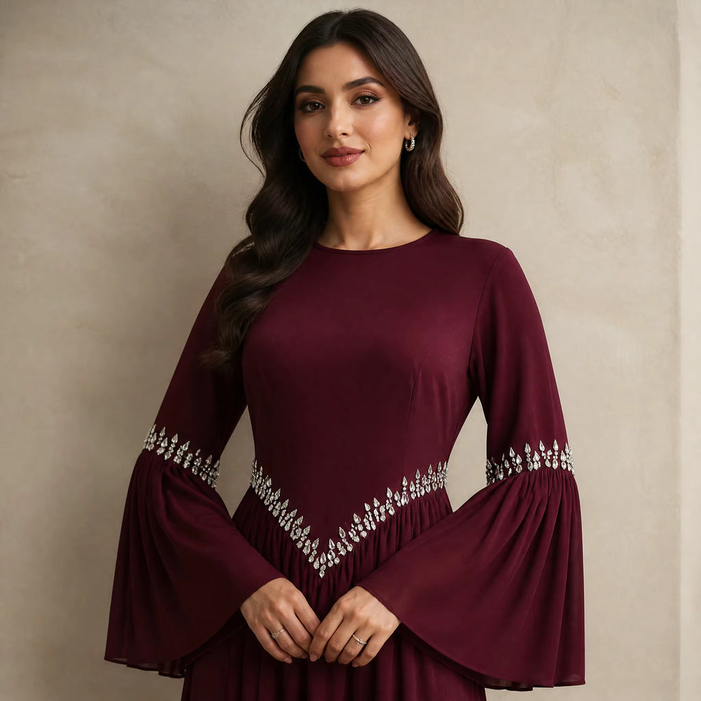 A woman wearing a long-sleeved burgundy dress with silver embellishments on the sleeves and waist, standing against a textured beige wall.