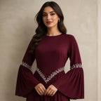 A woman wearing a long-sleeved burgundy dress with silver embellishments on the sleeves and waist, standing against a textured beige wall.
