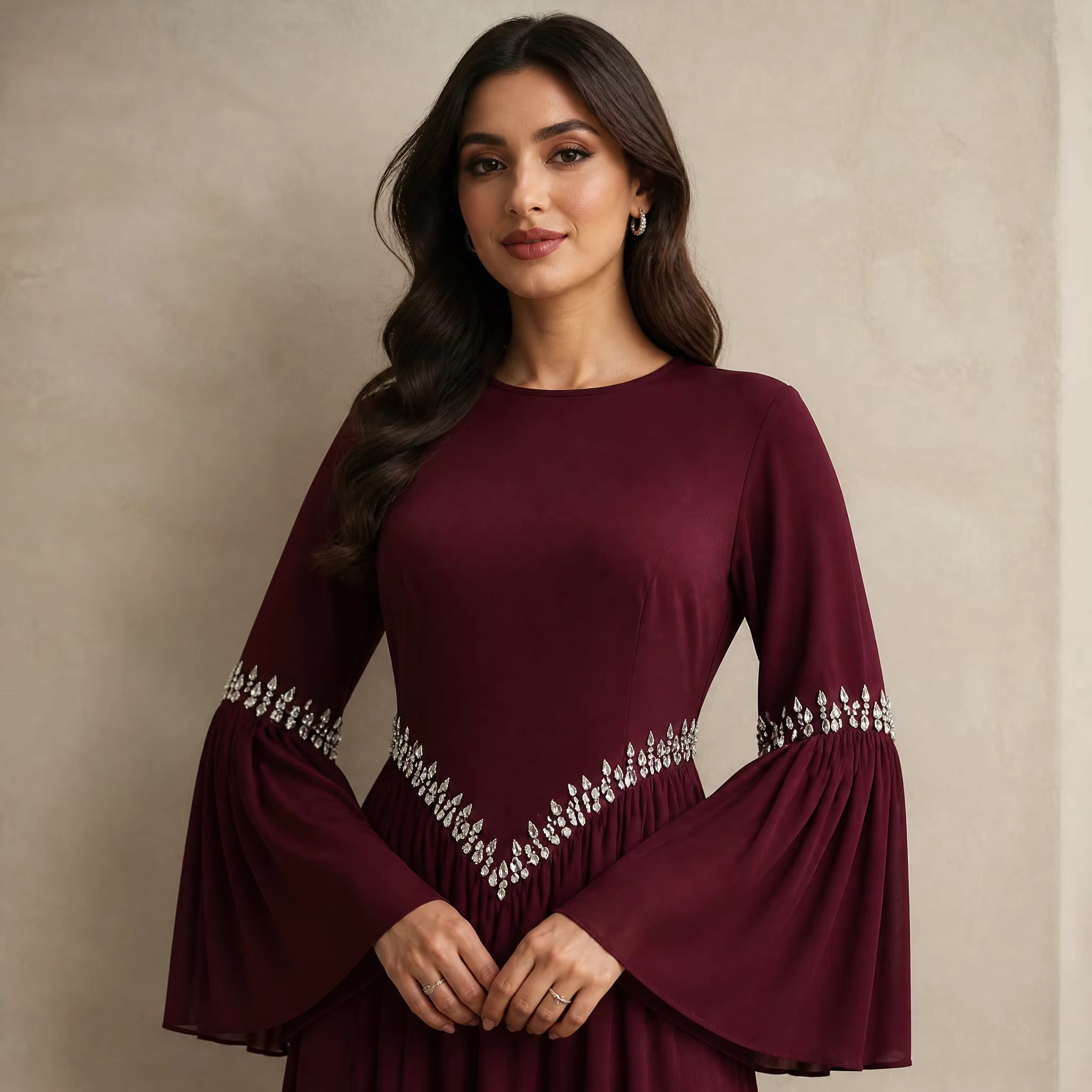 A woman wearing a long-sleeved burgundy dress with silver embellishments on the sleeves and waist, standing against a textured beige wall.