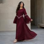 A woman modeling a long burgundy maxi dress with dramatic bell sleeves and silver beaded detailing at the waist and sleeve hems, standing in a modern stone walkway.