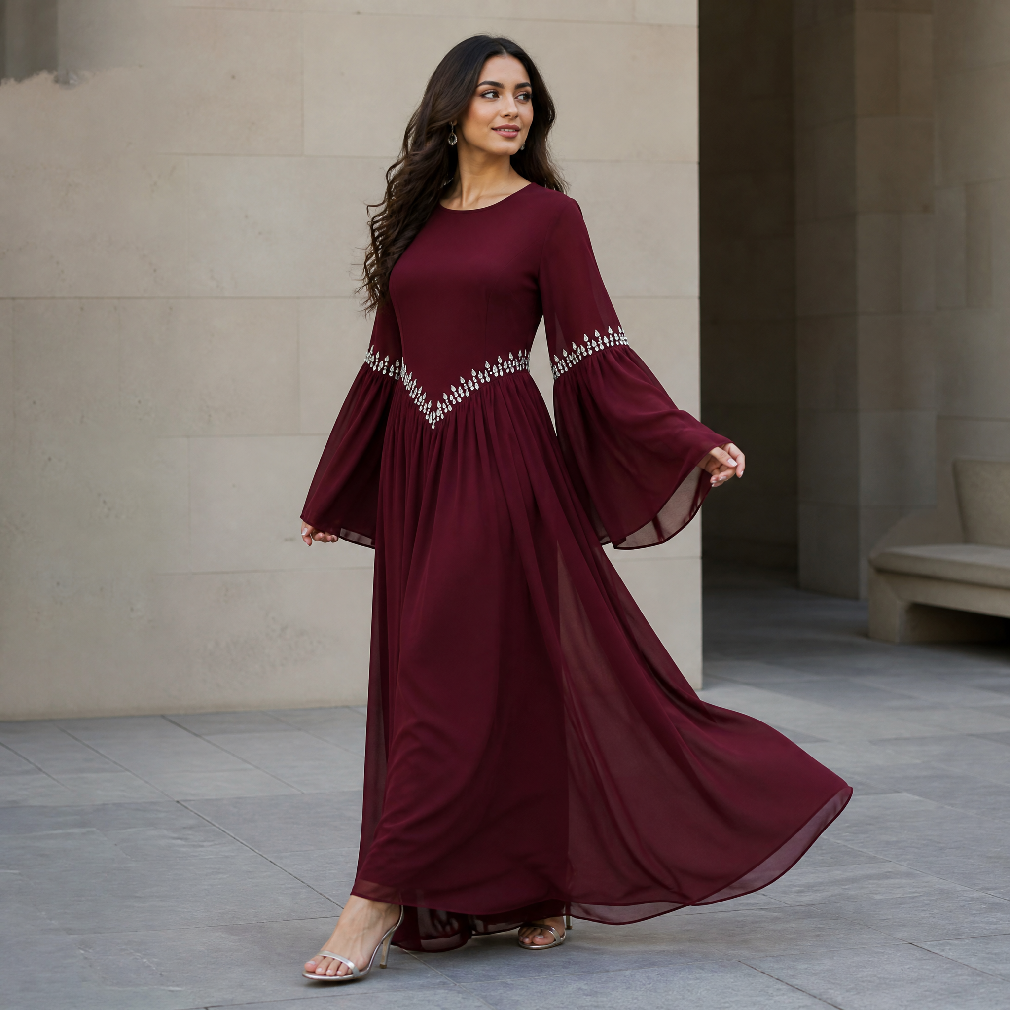 A woman modeling a long burgundy maxi dress with dramatic bell sleeves and silver beaded detailing at the waist and sleeve hems, standing in a modern stone walkway.