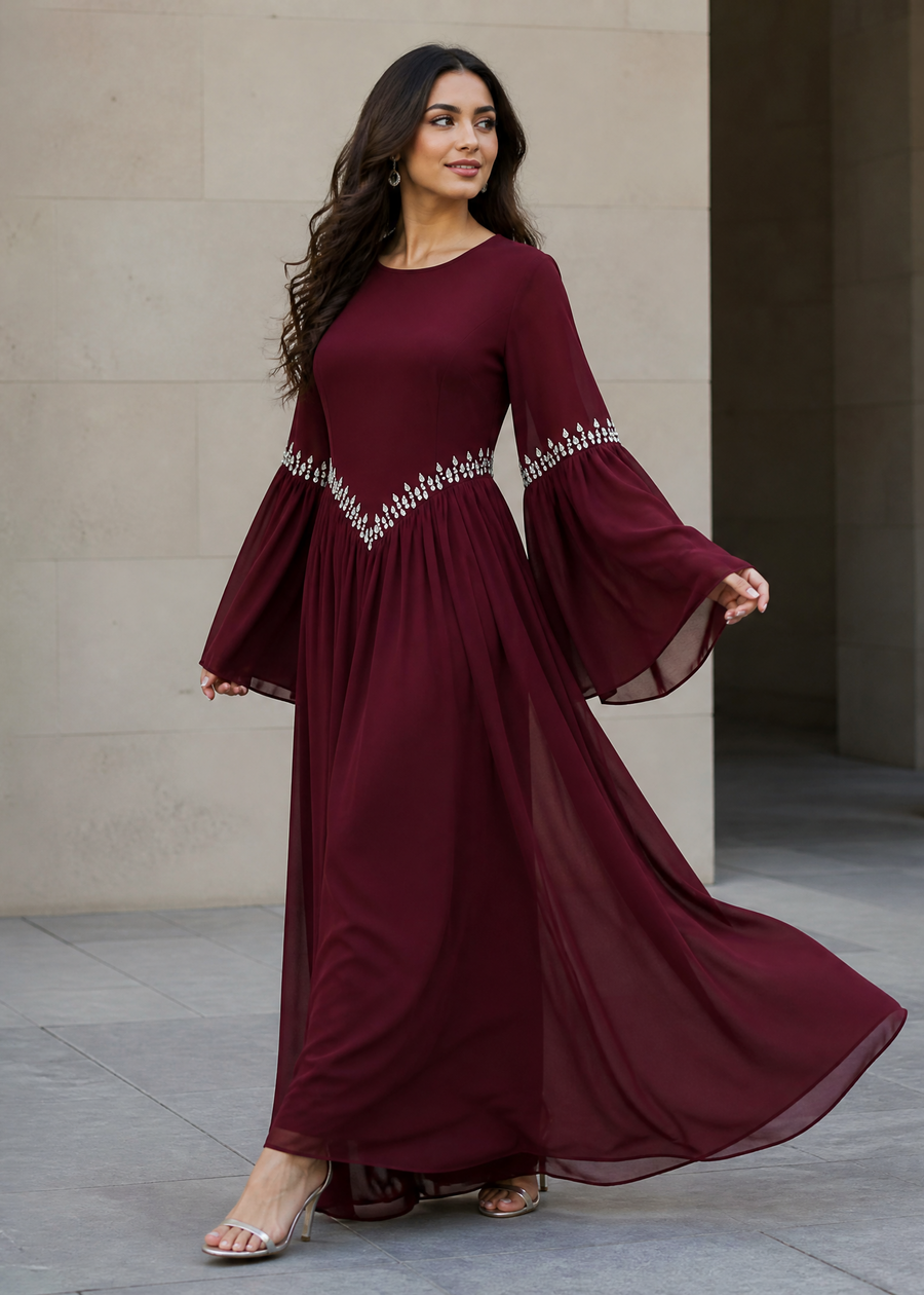 A woman modeling a long burgundy maxi dress with dramatic bell sleeves and silver beaded detailing at the waist and sleeve hems, standing in a modern stone walkway.
