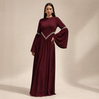 A woman modeling a floor-length burgundy maxi dress with long flared sleeves and silver beaded trim at the waist and sleeve cuffs, photographed against a plain beige studio backdrop.