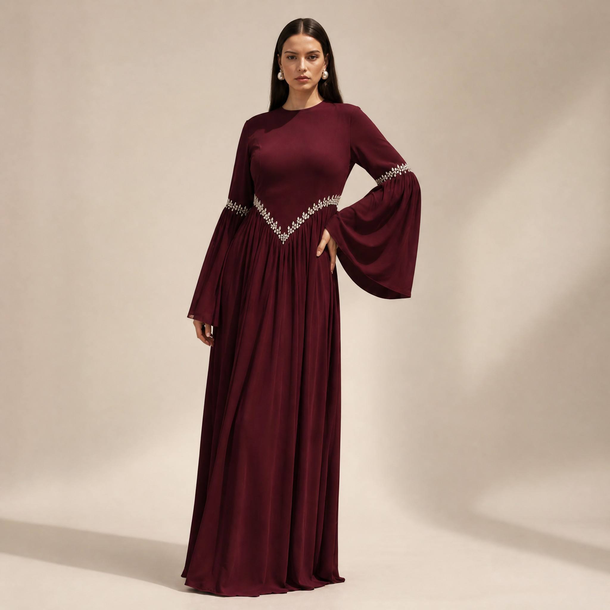 A woman modeling a floor-length burgundy maxi dress with long flared sleeves and silver beaded trim at the waist and sleeve cuffs, photographed against a plain beige studio backdrop.