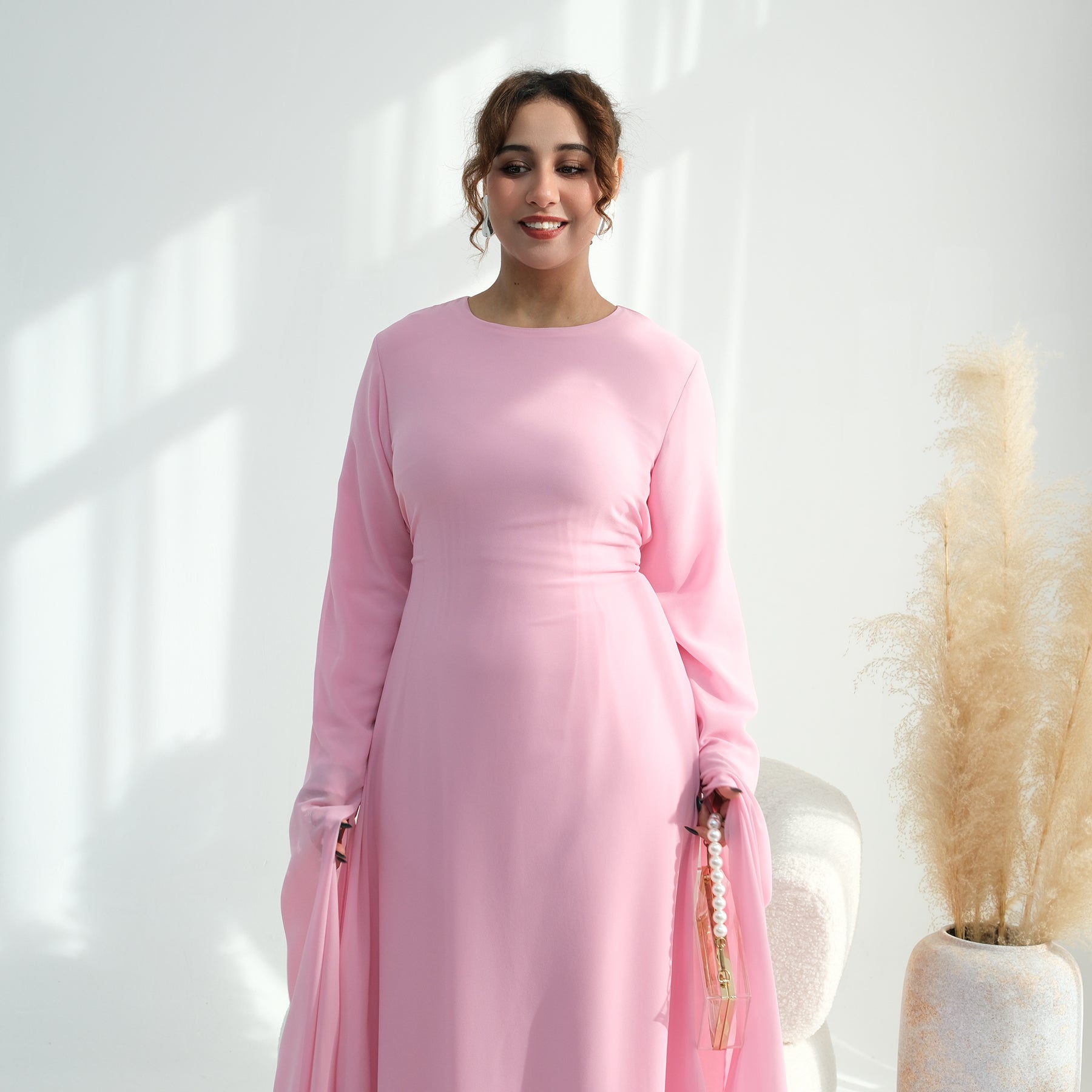 Woman wearing a pink dress standing in a bright room with a plant in the background