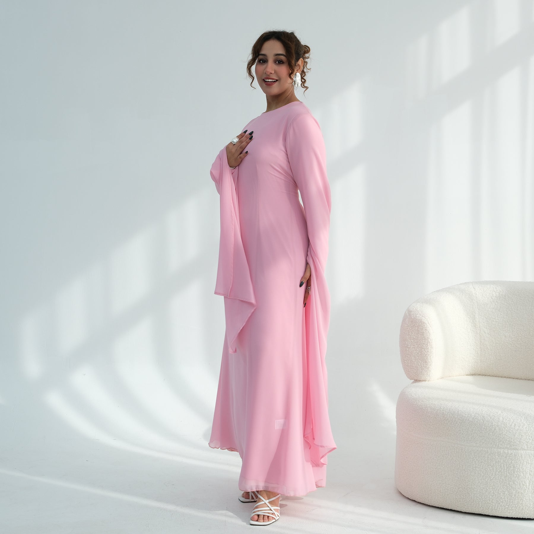 Woman in a pink outfit standing in a softly lit room with a white chair.