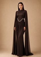 A woman modeling a floor-length dark brown gown with long draped cape sleeves, a high mock neck, and a wavy silver embellishment at the waist, set against a solid beige studio backdrop.