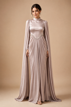 A woman modeling a floor-length satin gown in a dusty blush pink color, featuring a mock neck, long draped cape sleeves, and a beaded waistband, set against a solid beige studio background.