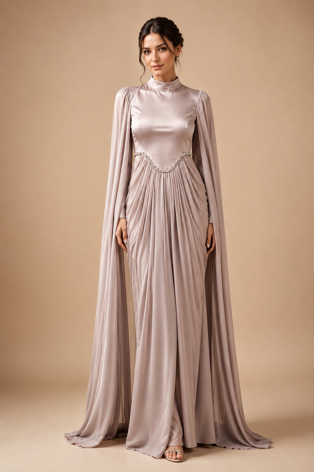 A woman modeling a floor-length satin gown in a dusty blush pink color, featuring a mock neck, long draped cape sleeves, and a beaded waistband, set against a solid beige studio background.