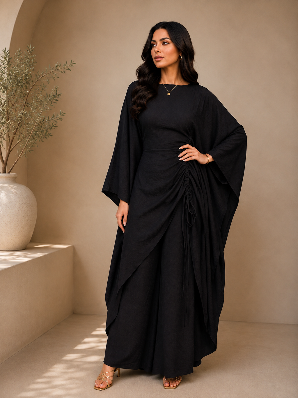 A woman modeling a black maxi dress with wide draped sleeves and a gathered, draped skirt, standing in a warm beige interior setting.