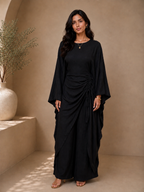 A woman modeling a black draped maxi dress with wide sleeves and a gathered waist tie, standing in a warm beige studio setting with a large vase of dried branches on the left.