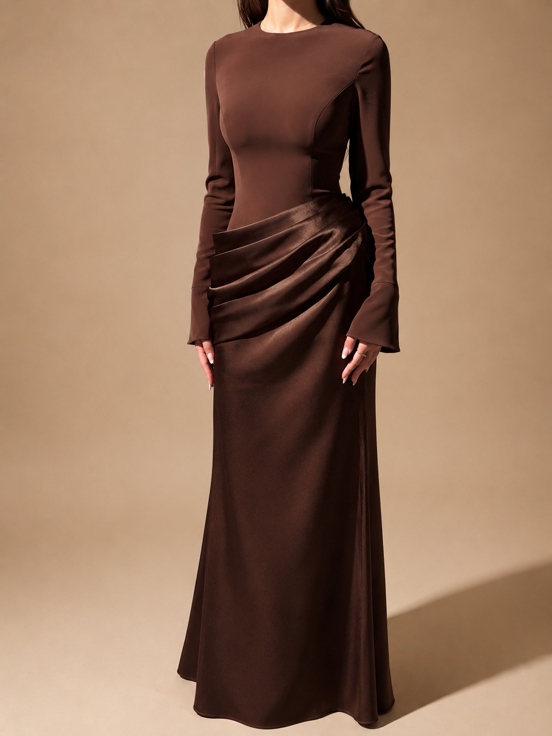 A woman wearing a long-sleeve, floor-length brown dress with a draped gathered waist, photographed against a plain warm beige studio background.