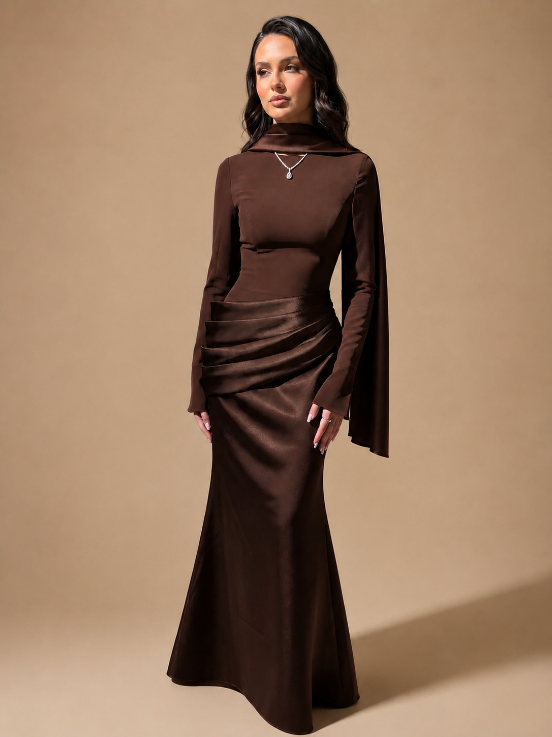 A woman modeling a dark brown long-sleeve mock-neck maxi dress with draped ruching and a flared skirt, photographed against a solid warm beige studio background.