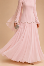 A woman wearing a flowing light pink long-sleeve maxi dress with sheer flutter sleeves, a scalloped embellished waist, and a full pleated skirt, holding a silver clutch against a warm beige background.
