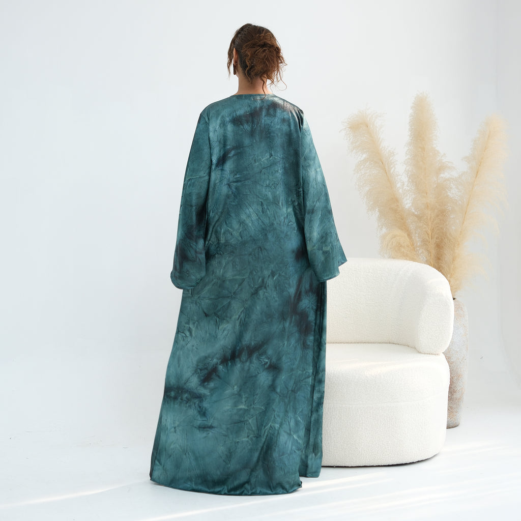 Person wearing a long teal dress standing in a minimalistic room with a white chair and potted plant.