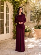 A woman wearing a floor-length, deep burgundy maxi dress with draped long sleeves and a gathered waist, standing in a sunlit stone courtyard with greenery and white-framed doors.