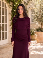 A woman wearing a long-sleeved, floor-length draped gown in deep burgundy, standing in a sunlit outdoor courtyard with stone walls, greenery, and large potted plants.