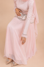 A woman wearing a long-sleeved light pink maxi dress with silver rhinestone embellishments on the cuffs and waist, holding a matching silver rhinestone clutch bag, set against a solid beige background.