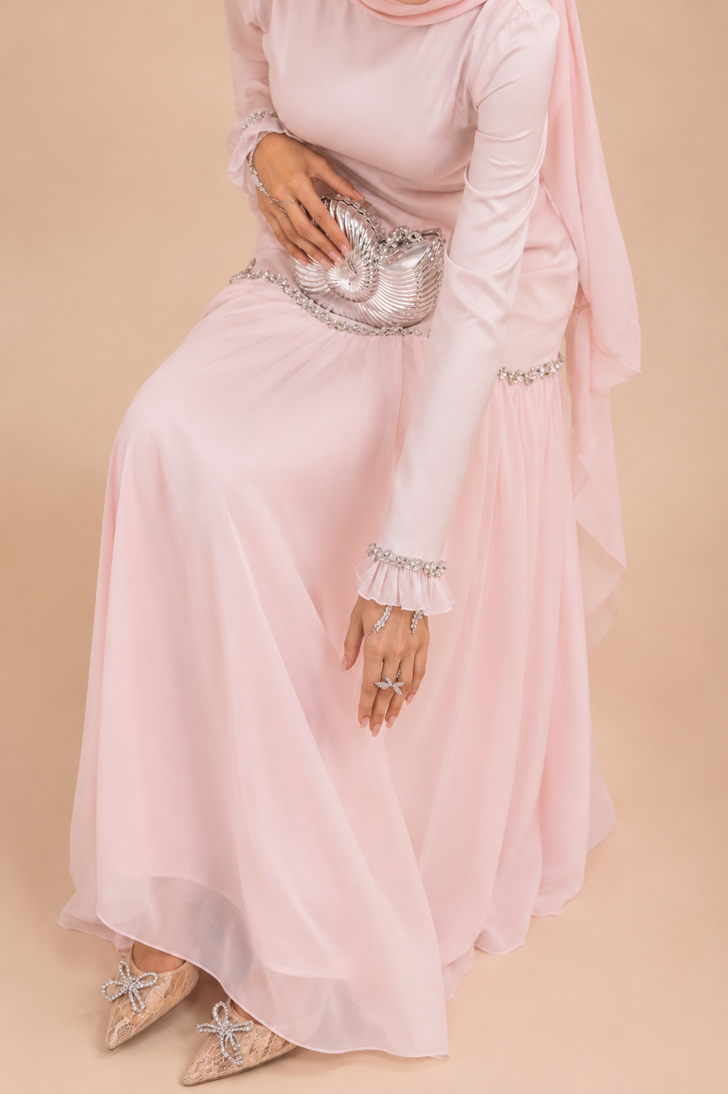 A woman wearing a long-sleeved light pink maxi dress with silver rhinestone embellishments on the cuffs and waist, holding a matching silver rhinestone clutch bag, set against a solid beige background.