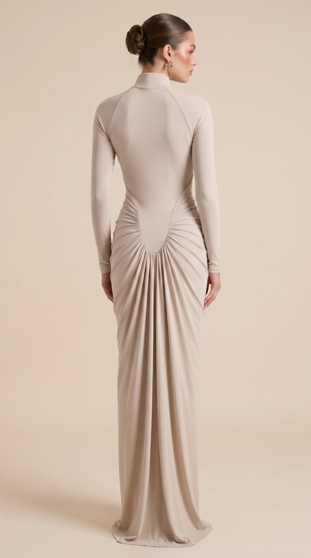 Woman wearing a long, beige dress with a high neckline and ruched back on a beige background