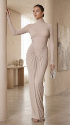 Woman in a beige long-sleeve gown standing in a softly lit room.