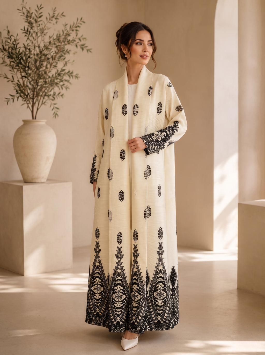 A woman models a long cream-colored abaya with black embroidered patterns, standing in a minimalist beige interior with a large vase of greenery on the left.