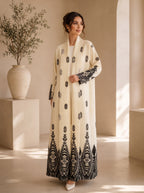 A woman models a long cream-colored abaya with black embroidered patterns, standing in a minimalist beige interior with a large vase of greenery on the left.