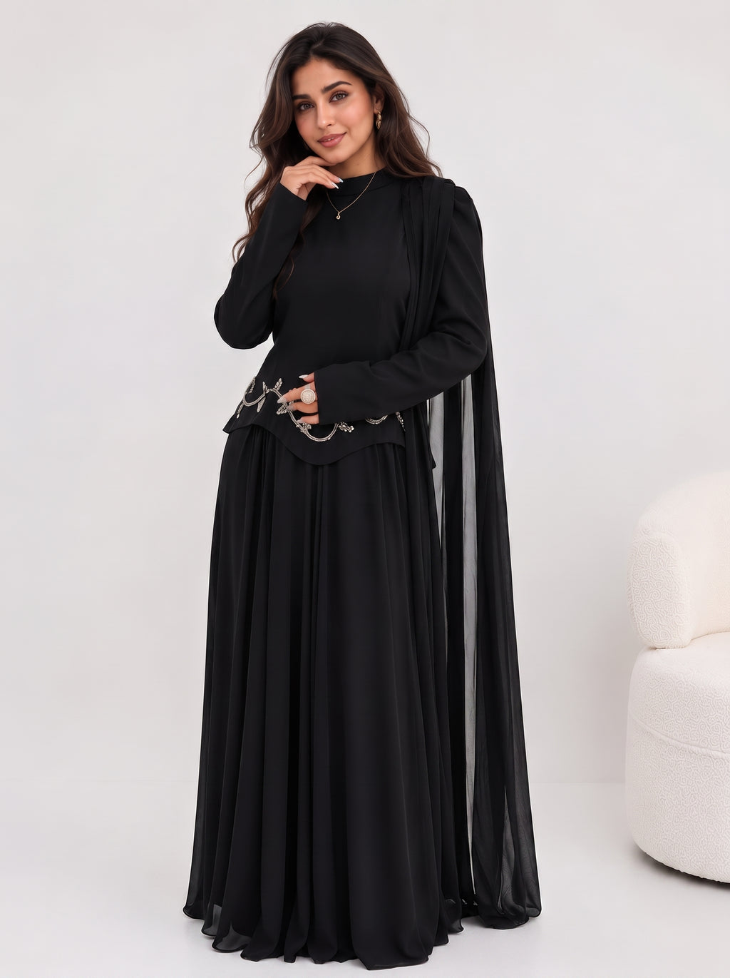 A woman modeling a long black dress with a sheer draped cape-like overlay, standing against a light gray studio background. A small cream upholstered chair is partially visible on the right side.