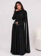 A woman modeling a long black dress with a sheer draped cape-like overlay, standing against a light gray studio background. A small cream upholstered chair is partially visible on the right side.