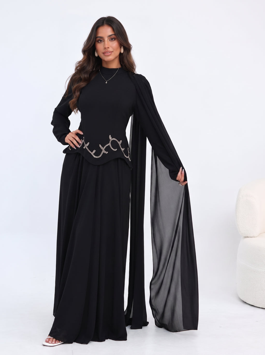 A woman modeling a black long-sleeve maxi dress with a sheer draped overlay and a decorative metallic chain detail at the waist, set against a clean white studio background.