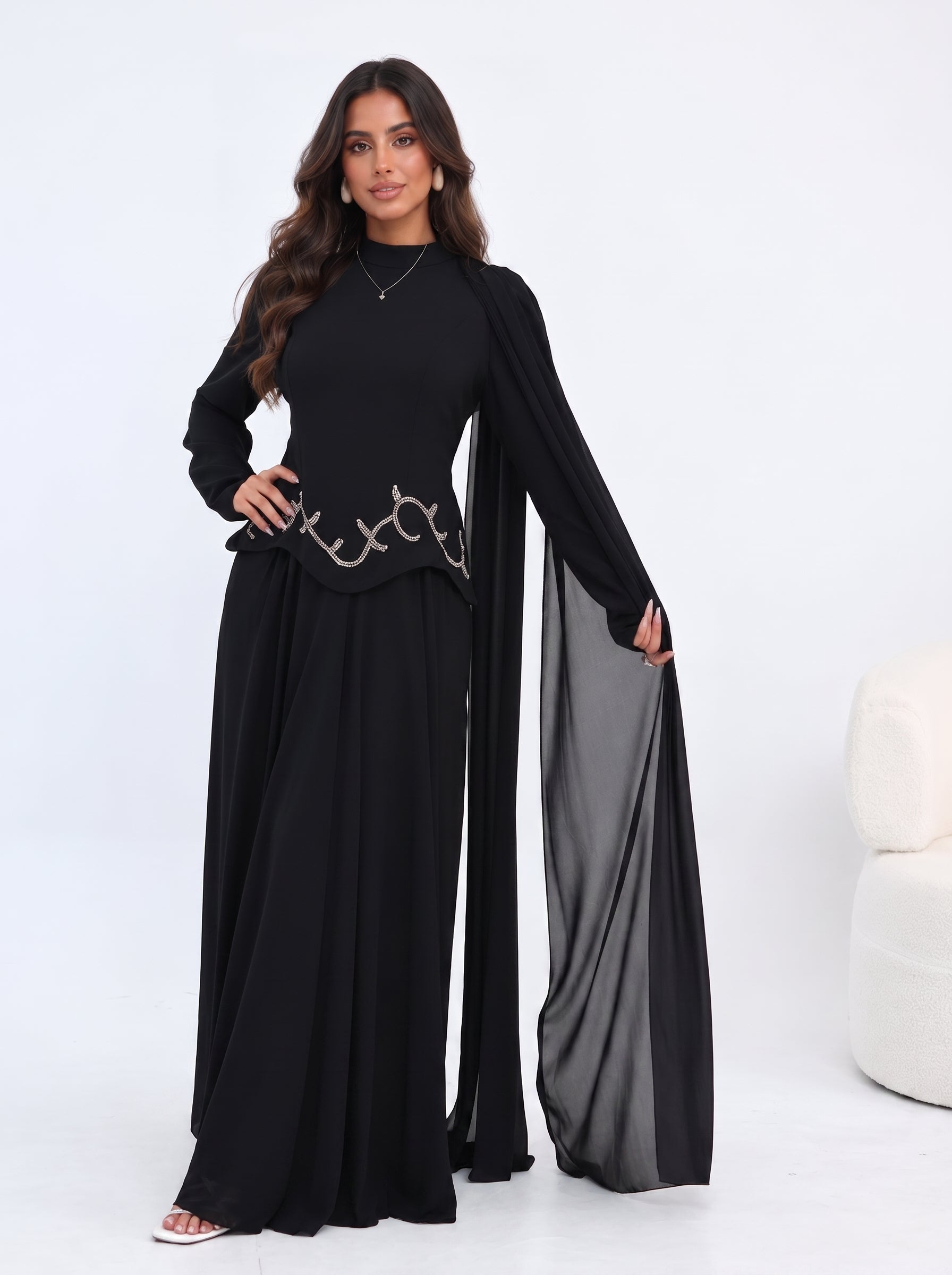 A woman modeling a black long-sleeve maxi dress with a sheer draped overlay and a decorative metallic chain detail at the waist, set against a clean white studio background.