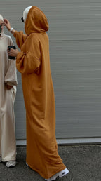 Person wearing a long mustard yellow hoodie with another person in a light-colored outfit standing next to them against a gray wall.