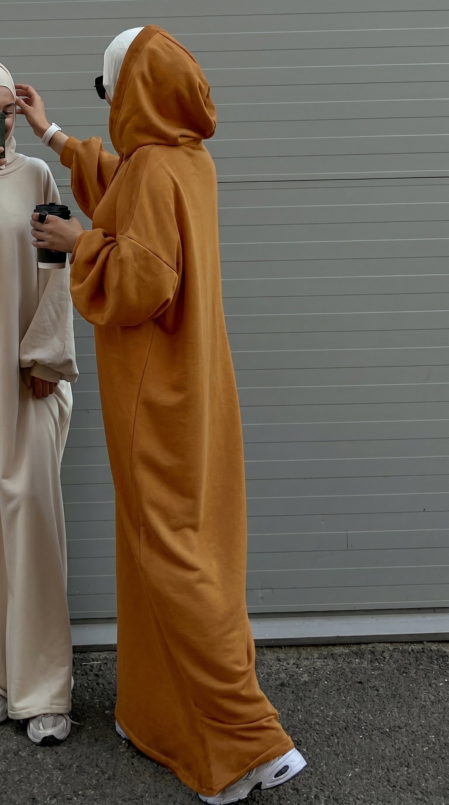 Person wearing a long mustard yellow hoodie with another person in a light-colored outfit standing next to them against a gray wall.