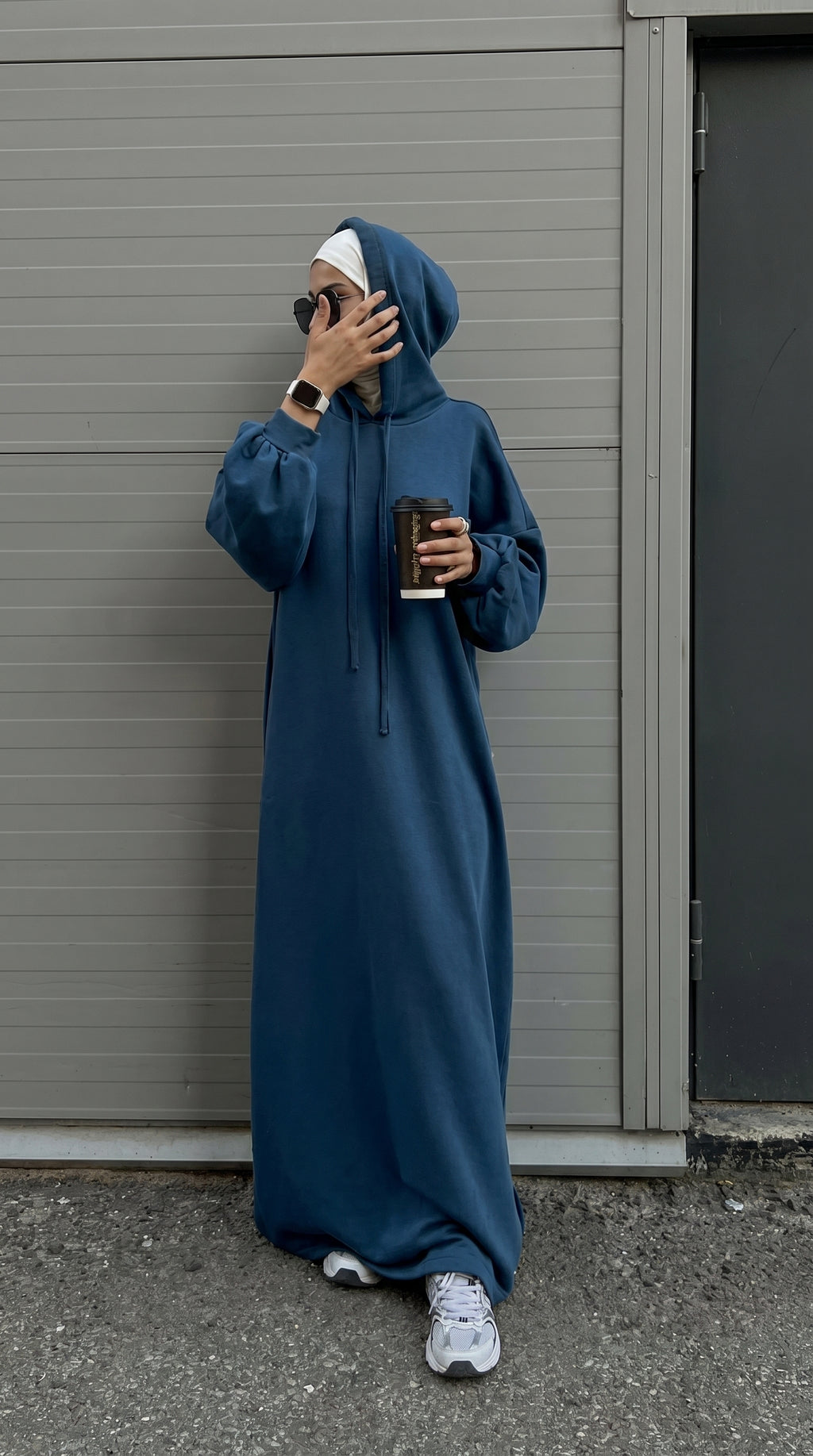 Person wearing a long blue hoodie standing against a gray wall.