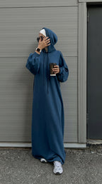 Person wearing a long blue hoodie standing against a gray wall.