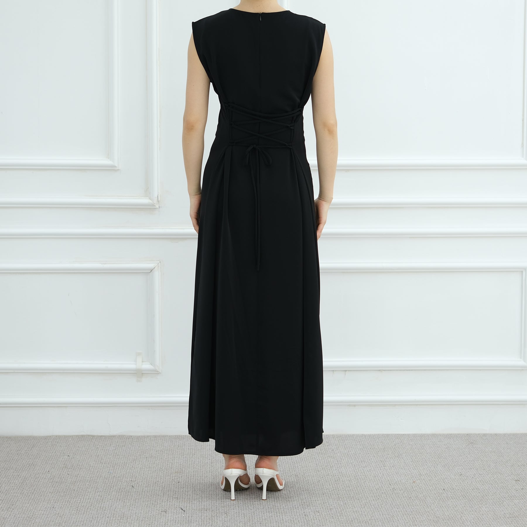 Person wearing a black sleeveless dress against a white paneled wall.