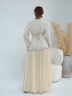 A woman viewed from the back wearing a long cream maxi dress with long sleeves, a lace-up back, and a pleated skirt. She stands in a bright studio setting with a white chair and pampas grass in a vase beside her.