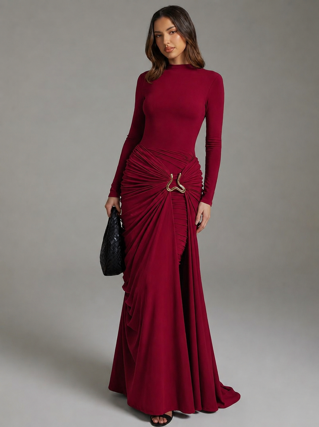A woman modeling a long-sleeve, floor-length burgundy maxi dress with a ruched draped skirt and a small metallic ring detail at the waist, holding a black clutch against a neutral gray studio background.