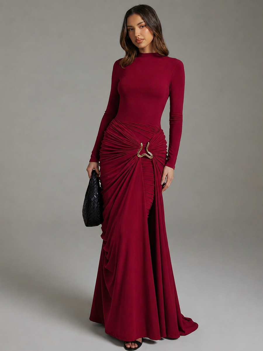 A woman modeling a long-sleeve, floor-length burgundy maxi dress with a ruched draped skirt and a small metallic ring detail at the waist, holding a black clutch against a neutral gray studio background.